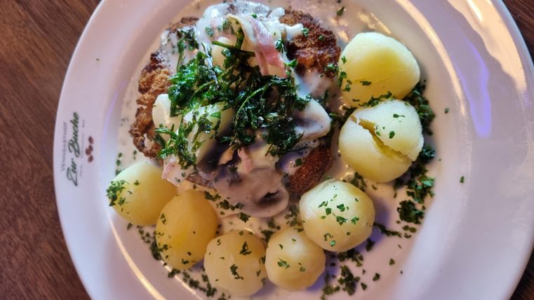 A delicious dish with a schnitzel-like main part, covered with a white sauce and fresh herbs. Served with neatly arranged boiled potatoes.
