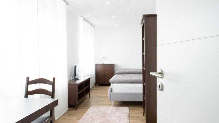 A bright, modern room with two beds and a small table. The walls are in neutral colors, and large windows provide plenty of daylight.