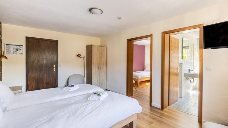 A bright room with two single beds and modern furniture. In the background, there is an open door to a bathroom and another sleeping area.