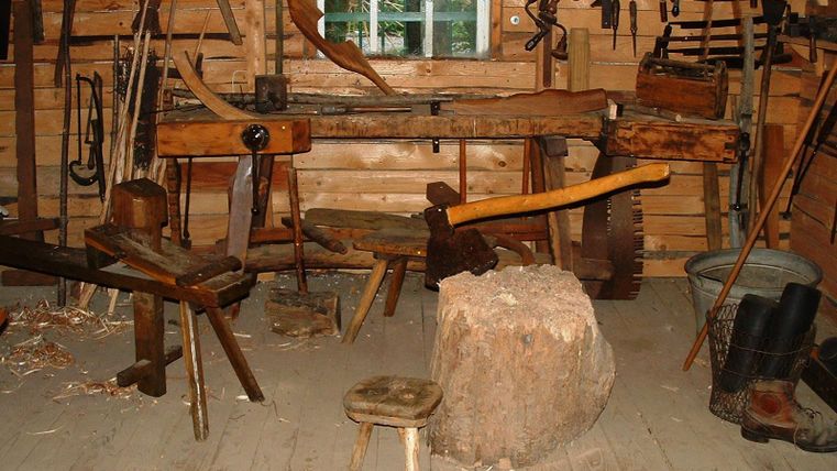 A traditional carpentry shop with various tools and woodworks. The space is rustic and made of wood, with a workbench in the center.