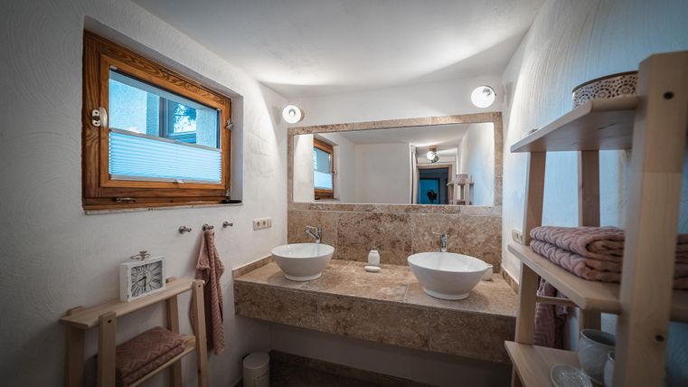 Een moderne badkamer met twee wastafels en een grote spiegel. De muren zijn licht en er zijn houten planken met handdoeken.