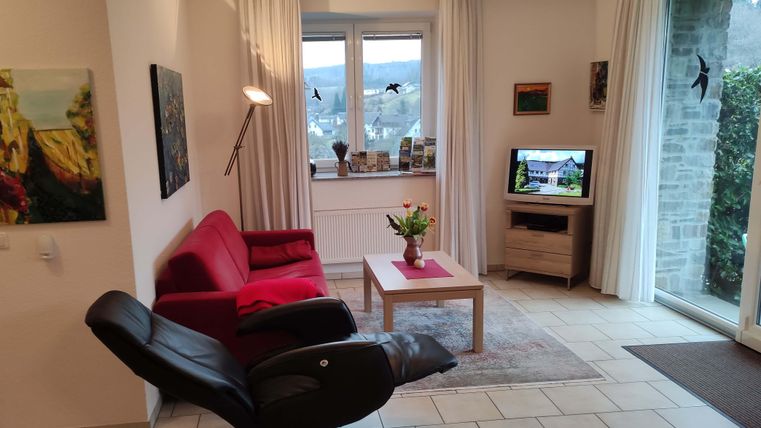 A cozy living room with a red sofa and a relaxing armchair. A table with a flower vase and a television are also visible.
