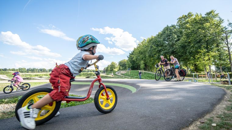 A small child is riding a balance bike on a paved path. In the background, two adults can be seen on bicycles.