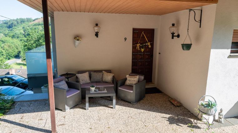 A cozy outdoor area with seating and a covered entrance. There are green plants in the background and a peaceful view.