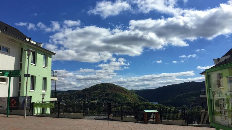 A picturesque view of gentle hills under a clear sky with a few clouds. In the foreground, green buildings and a terrace are visible.