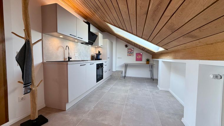 Een moderne keuken onder het dak met houten plafonds en een lichte vloer. Er is een royale raamoppervlakte die veel licht binnenlaat.