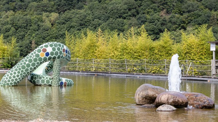 Een kleurrijke speeltuin met een golvende glijbaan en een fontein in een rustig waterbassin. Op de achtergrond zijn groene bomen en heuvels zichtbaar.