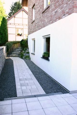 A narrow path runs along a house's white-painted wall, with a paved and gravel-covered area. In the background, some plants and another building are visible.