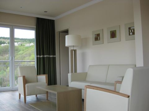 A modern living room with a white sofa and wooden chairs. Large windows offer a view of the green countryside.