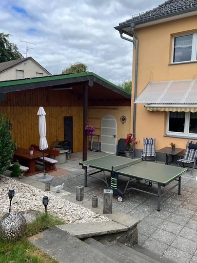 A cozy patio with a table tennis court and a wooden pavilion. The area is surrounded by plants and a bright house.