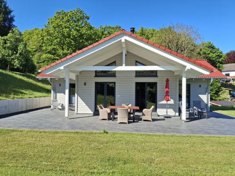 Een modern huis met een rood dak en veranda. De tuin is groen en goed onderhouden, omringd door bomen.