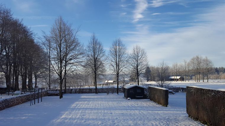 Een winterlandschap met besneeuwde bomen en een heldere lucht. Op de achtergrond zijn kleine hutten en velden zichtbaar.