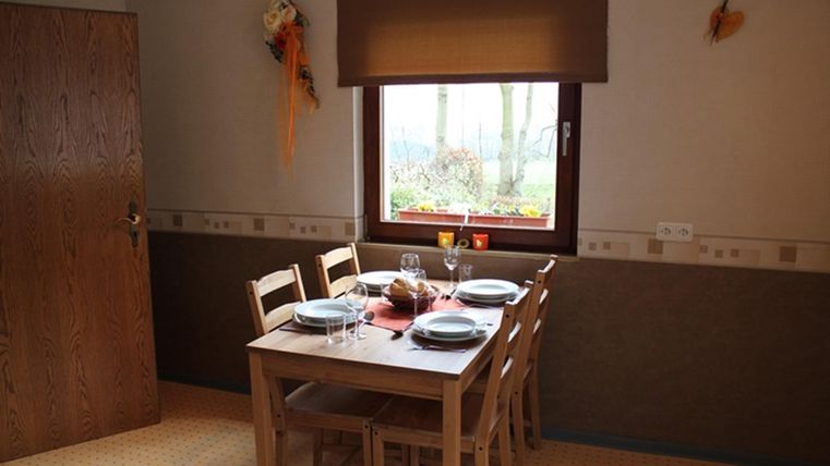 A simple dining area with a wooden table and chairs. Placed on the table are set plates and glasses, and a window lets in light.