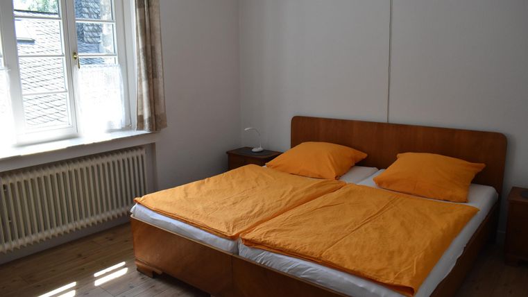 Ein einfaches Schlafzimmer mit einem Doppelbett und orangefarbenen Kissen. Natürliches Licht strömt durch das Fenster.