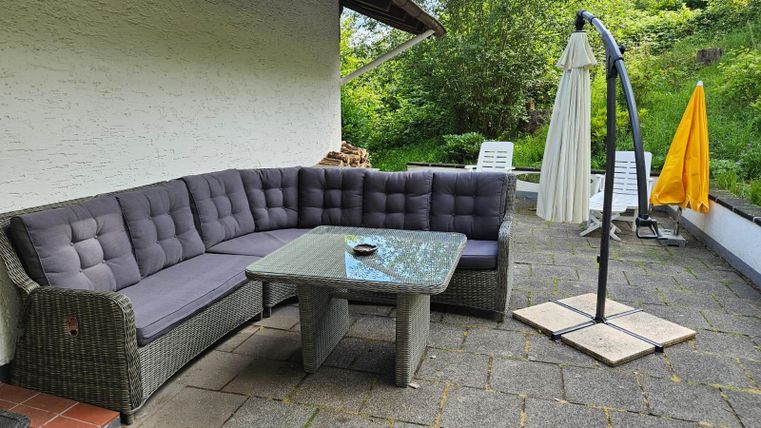 Eine gemütliche Sitzecke mit einer grauen Rattan-Sofagarnitur und einem Glastisch. Im Hintergrund sind grüne Pflanzen und ein Sonnenschirm sichtbar.
