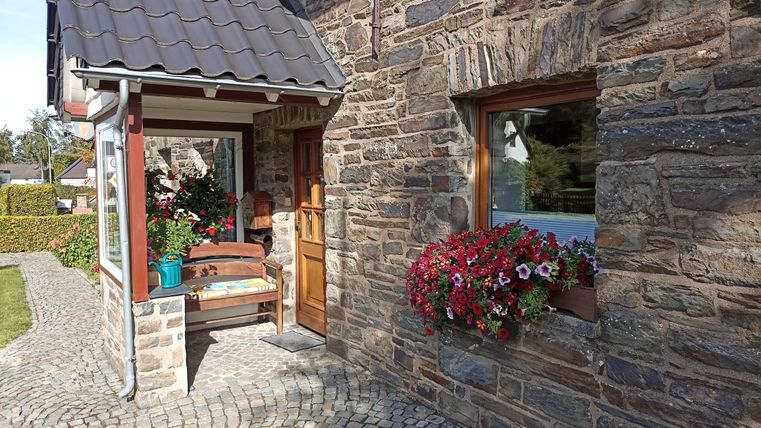 Ein einladender Hauseingang mit Steinmauern und einem bepflanzten Fenster. Blumen in einem Topf schmücken den Eingangsbereich und sorgen für eine freundliche Atmosphäre.