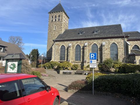 Een oude kerk met een markante toren staat op de voorgrond. Op de voorgrond staat een rode auto en rondom zijn verzorgde planten en een parkeerzone te zien.