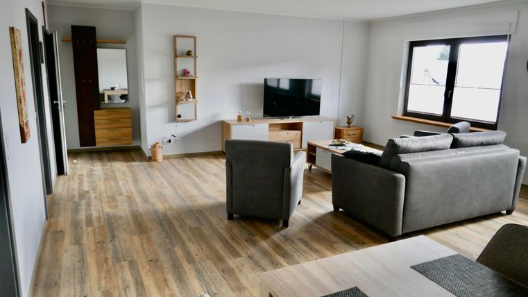 A modern living room with a gray sofa and a wooden table. The room is bright with large windows and a television on the wall.