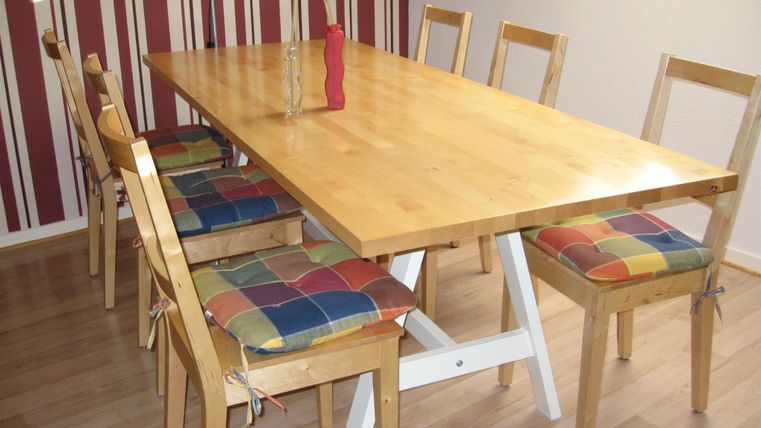 Een eenvoudige eettafel van hout met kleurrijke stoelen. Op de achtergrond zijn gestreepte muren te zien.
