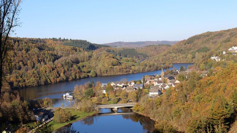 Eine malerische Landschaft mit einem Fluss, umgeben von bunten Herbstbäumen. Im Hintergrund sind kleine Häuser und ein Brücke zu sehen.