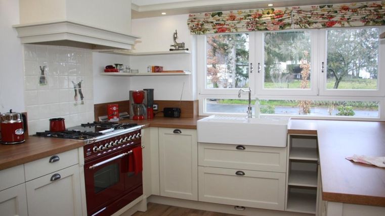 A modern kitchen with white cabinets and a red stove. Large windows provide plenty of daylight and a view of the outdoors.