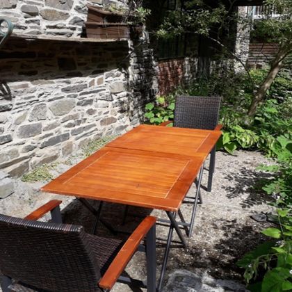 A wooden table with two chairs is located in a green outdoor area. In the background, a stone wall and plants can be seen.