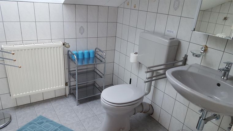 Een badkamer met witte tegels, een wastafel en een toilet. Er is ook een handdoekhouder en een kleine opbergruimte voor toiletartikelen.