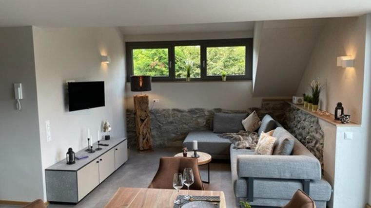 Een moderne woonkamer met een gezellige bank en een houten tafel. Grote ramen laten veel licht binnen en bieden uitzicht op de natuur buiten.