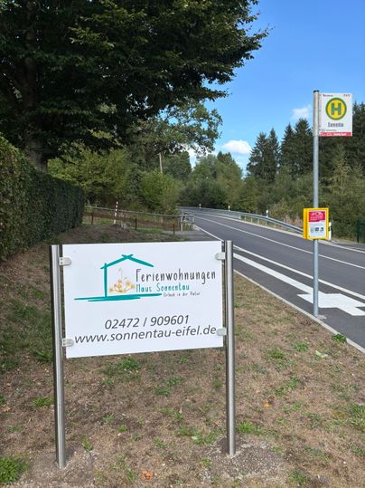Een wegwijzer naar vakantieappartementen genaamd "Haus Sonnentau" staat aan een straat. Naast de wegwijzer bevindt zich een bushalte in een landelijke omgeving.