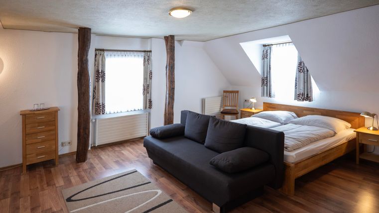 A cozy bedroom with wooden flooring and bright light. There is a large bed, a sofa, and windows with curtains.