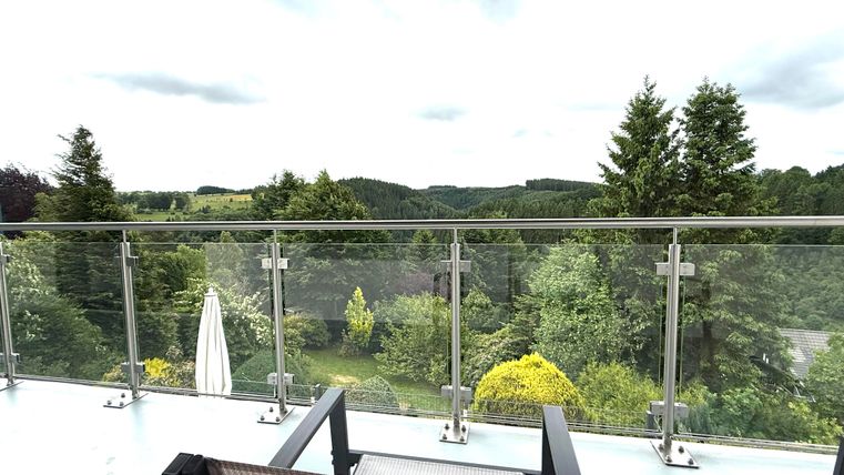 Een balkon met uitzicht op een groen landschap. De omgeving is gekenmerkt door bomen en heuvels onder een bewolkte lucht.
