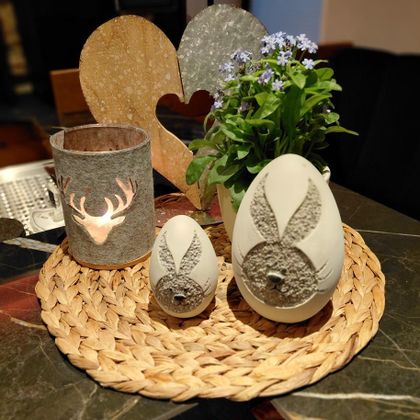 A decorative arrangement of spring accessories on a table. There are two eggs with bunny motifs, a small container with a deer motif, and a flowering plant in a pot.