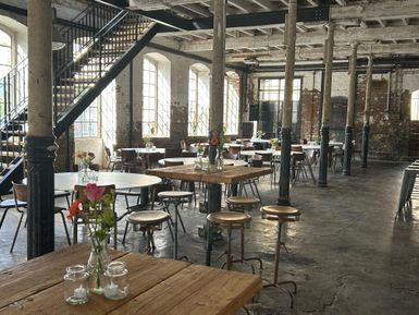 Ein stilvolles Restaurant mit offenen Räumen und hohen Fenstern. Die Einrichtung ist modern und rustikal, mit Holztischen und Metallstühlen.