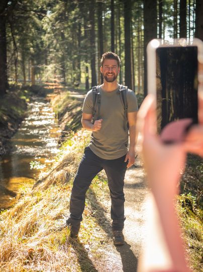 Een glimlachende man staat op een bosweg naast een kleine beek. Hij houdt een telefoon in zijn hand en poseert voor een foto.
