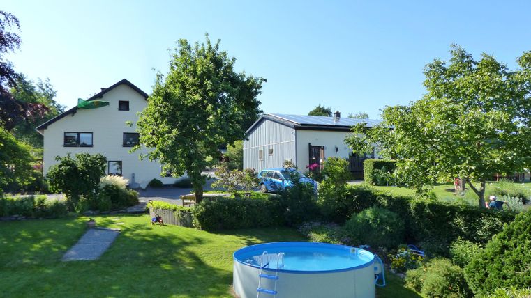 A beautiful garden with a blue pool and plenty of greenery. In the background, there are two houses and trees visible.