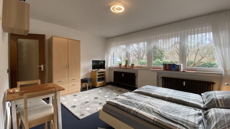 A cozy bedroom with two beds and a desk. Large windows let in plenty of daylight.
