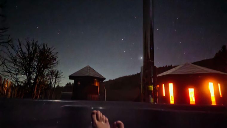 Een ontspannend bad in de buitenlucht onder een heldere sterrenhemel. Op de achtergrond zijn twee verlichte gebouwen en bomen zichtbaar.
