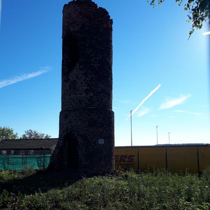 An old, broken tower made of stone stands in a green meadow. In the background, a blue sky and some clouds can be seen.