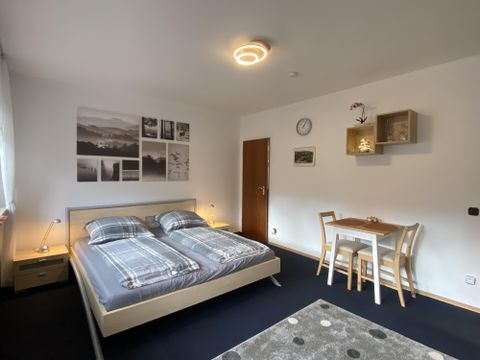 A modern bedroom with a double bed and wooden cabinets. A small table with chairs and decorative pictures on the wall give the room a cozy atmosphere.
