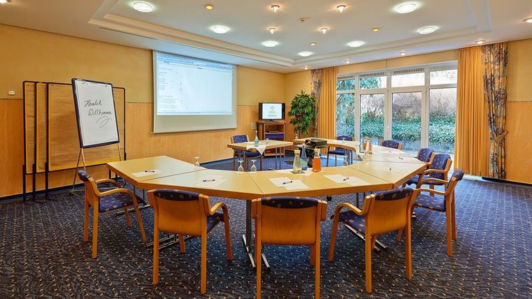 Ein moderner Konferenzraum mit einem großen, ovalen Tisch und Stühlen. Es gibt ein Whiteboard, einen Bildschirm und viel Tageslicht durch die Fenster.