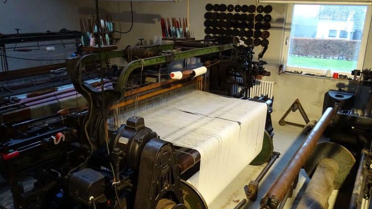 An old weaving machine in a workshop-like room. The table is covered with a large piece of woven fabric and provides insight into textile production.