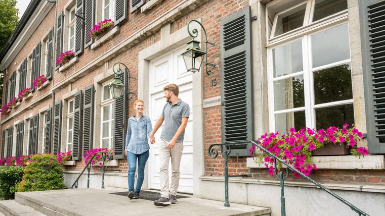 A charming building with windows decorated with green shutters and colorful flowers. Two people are standing on the steps and smiling.