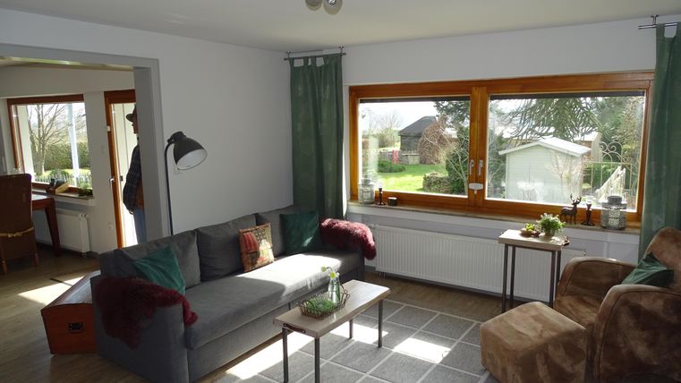 Een gezellige woonkamer met een grijze bank en een beige fauteuil. Grote ramen bieden uitzicht op de tuin.