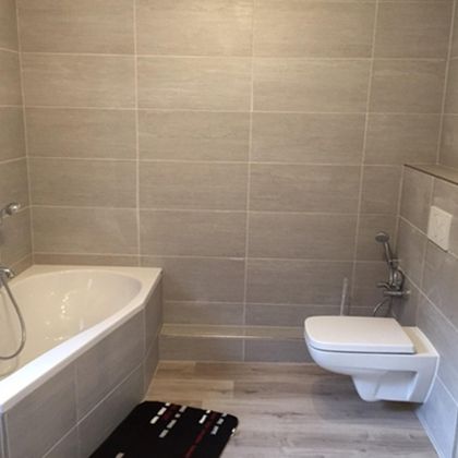 A modern bathroom with a bathtub and a toilet. The walls are clad with gray tiles and the floor is wooden.