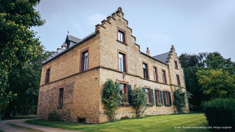 Een historisch stenen huis met puntdaken en nostalgische charme. Omringd door een groene tuin en bomen.