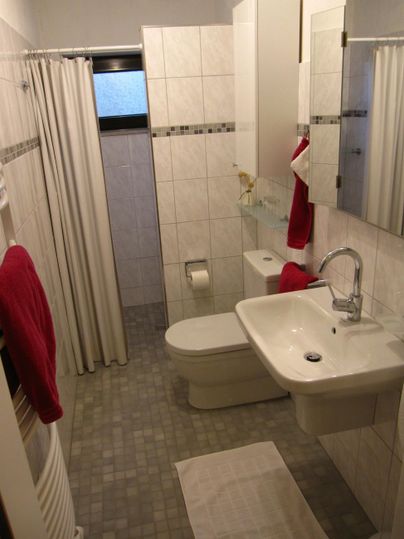 Ein kleines Badezimmer mit weißen Fliesen und einer Duschkabine. Es gibt ein Waschbecken, eine Toilette und Handtücher in Rot.