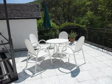 Een gezellige balkon met een ronde tafel en vier witte stoelen. Op de achtergrond is een groene parasol en veel groen loof te zien.