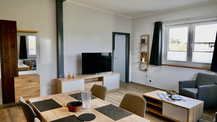A modern living room with a large television, a dining table, and a cozy couch. The walls are bright, and the windows provide plenty of daylight.