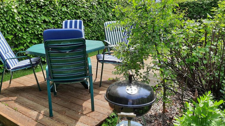 A cozy dining area in the garden with a round table and chairs. Next to the table is a grill, and the surroundings are green and planted.