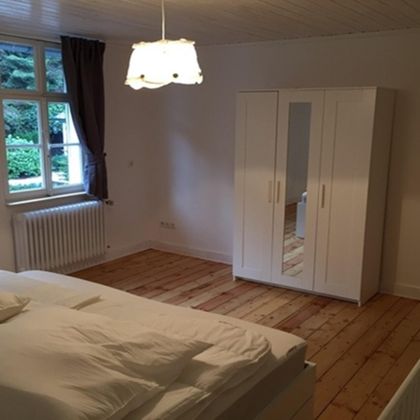 A simple bedroom with a large bed and wooden floor. A bright wardrobe and a window with curtains create a pleasant atmosphere.